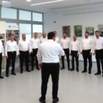 Moški pevski zbor v belem stoji v polkrogu in nastopa v svetlem notranjem prostoru doma starejših. Dirigent stoji s hrbtom proti kameri in vodi zbor. V ozadju so knjige, umetniške slike in ura na steni.
