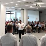 Moški pevski zbor nastopa pred številnim občinstvom v domu starejših. Dirigent stoji pred pevci in usmerja petje. Občinstvo, sestavljeno iz starejših oseb, z zanimanjem spremlja nastop.