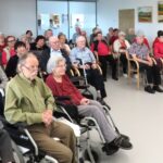Občinstvo v domu starejših z zanimanjem spremlja pevski nastop. Večina obiskovalcev sedi, nekateri so v invalidskih vozičkih ali uporabljajo pripomočke za hojo. V ozadju so umetniške slike na steni.