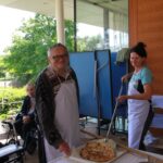 Starejši moški in ženska v predpasnikih pripravljata pico na zunanjem prostoru doma starejših, v ozadju sedijo stanovalci z vozički.