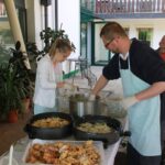 Moški in ženska pripravljata ocvrte bezgove cvetove na stojnicah doma za starejše, v ozadju je kuhinjska oprema in čakalna vrsta.