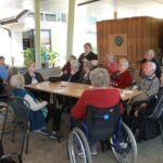 Skupina stanovalcev doma za starejše, večina v invalidskih vozičkih, pozorno posluša žensko, ki govori ob leseni mizi v skupnem prostoru doma.