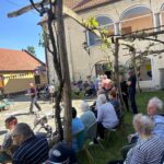 Starejši obiskovalci sedijo na stolih na dvorišču pred starim grajskim poslopjem. Nekateri se pogovarjajo, drugi opazujejo dogajanje. V ozadju so okrašene stavbe in ljudje v druženju. V ospredju moški z očali gleda proti kameri.
