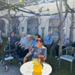 Skupina starejših oseb sedi v senci pod pergolo z vinsko trto pred grajsko stavbo. Nekateri imajo hojce ali invalidske vozičke. Na mizici je vrč z osvežilno pijačo. V ozadju stoji nekaj oseb v pogovoru.