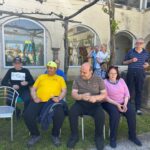 Starejši moški in ženske sedijo in stojijo ob grajski stavbi. Nekateri imajo v rokah kozarce ali skodelice. V ozadju so okna z odsevom napihljive igralne naprave, mimo hodijo drugi obiskovalci.