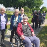 Skupina stanovalcev doma za starejše se sprehaja po urejenem vrtu. V ospredju gospa v invalidskem vozičku, za njo nekaj oseb z berglami in invalidskimi vozički. Sončen dan in veliko zelenja.