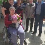 Skupina obiskovalcev pozira za skupinsko fotografijo. V ospredju gospa v invalidskem vozičku s šopkom cvetja, okoli nje še štiri osebe, vsi nasmejani.