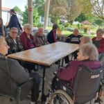 Stanovalci sedijo ob velikem lesenem mizu pod nadstreškom v senčnem vrtu. Okoli mize se družijo in klepetajo. V ozadju sta vidna urejen park in paviljon.