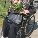 Starejša gospa v invalidskem vozičku drži šopek potonik in se nasmejana ozira proti kameri. V ozadju zelenica in ograja.