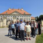 Skupina ljudi stoji pred zgodovinsko stavbo z baročnim pročeljem in rdečo streho, obdanimi z ograjo.
