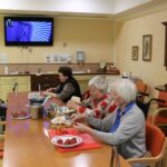 Več starejših oseb, od katerih so nekatere v invalidskih vozičkih, sedi ob dolgi mizi v skupnem prostoru doma za ostarele. Nekatere režejo jagode in pripravljajo sestavine. Na zidu visi televizor z modro-vijolično sliko, v ozadju je kuhinjski pult.