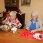 Dve starejši ženski sedita ob mizi in pripravljata sestavine – ena reže jagode na rdeči deski, druga dodaja sestavine v veliko kovinsko skledo. Na mizi so še druge posode in sestavine.