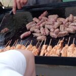 Priprava hrane na žaru: oseba peče čevapčiče in piščančje ražnjiče, ob strani so lonci s hrenovkami in sokovi.