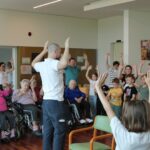 Starejši stanovalci doma in otroci skupaj izvajajo gibalno aktivnost. Moški v belem vodi vajo z dvignjenimi rokami, vsi udeleženci posnemajo gibanje in se zabavajo.