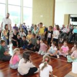 Skupina otrok se pripravlja na igro z žogicami, medtem ko starejši gledalci sedijo v ozadju. Prostor je svetel in prijeten, vzdušje sodelovalno.