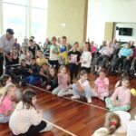 Otroci sodelujejo v gibalni igri z žogicami, medtem ko jih starejši stanovalci doma z zanimanjem opazujejo in spodbujajo. Dogodek poteka v večnamenskem prostoru.