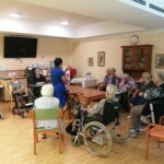 Skupina starejših oseb, večinoma žensk, sedi v krogu med šiviljsko delavnico. V ospredju stoji izvajalka v modri obleki in prikazuje ročno delo. Udeleženke pozorno spremljajo prikaz.