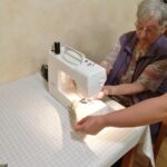 Starejša udeleženka šiviljske delavnice šiva kos blaga s pomočjo bele šivalne mašine. Osebje ji pomaga pri nastavitvi blaga.