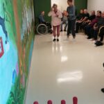 Stanovalka doma starejših s pomočjo zaposlene stoji pripravljena na met žoge pri igri kegljanja, ob strani sedijo drugi stanovalci kot občinstvo.