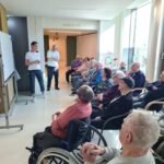 Več stanovalcev doma starejših sedi v polkrogu in pozorno spremlja predstavitev pred belo tablo, ki jo vodi zaposlena.