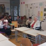 V učilnici s plakati na stenah skupina osnovnošolcev sedi v krogu z dvema starejšima osebama. Ena učenka igra harmoniko. V ozadju so razvidni šolski pripomočki, učna tabla in gasilni aparati. Vzdušje je sproščeno in vključujoče.