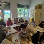 Otroci in starejši skupaj sedijo za mizami, okrašenimi z različno semensko dekoracijo. V ospredju učiteljica pomaga pri ustvarjanju. Na oknih so živobarvni papirnati metulji in rože, zunaj pa je viden otroški tobogan.