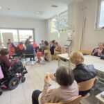 V svetli učilnici ob mizah sedijo starejši in mlajši udeleženci, nekateri v invalidskih vozičkih. Vsi izdelujejo papirnate rože. V ozadju je vidna okrasitev in naravna svetloba skozi okna.