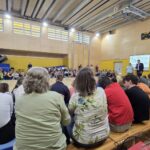V športni dvorani poteka prireditev z več deset udeleženci različnih starosti, ki sedijo v krogu. Dva govorca stojita ob platnu z napisom o svetovnem dnevu cerebralne paralize. Dogodek spremlja občinstvo s tribune.