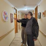 Starejši moški in mlajša ženska stojita na hodniku doma in gledata razstavljene slike na steni. Moški kaže na eno izmed umetniških del, oba se nasmihata.