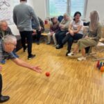 Skupina starejših in mlajših oseb se udeležuje družabne igre s keglji v sobi. Starejši moški v ospredju kleči in vrže rdečo kroglo proti pisanemu setu kegljev.