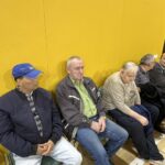 Skupina moških in žensk različnih starosti sedi ob rumeni steni telovadnice. Nekateri gledajo naravnost, drugi so v pogovoru. V ozadju stoji še nekaj oseb ob vratih.