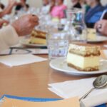 Na leseni mizi je v ospredju kos kremne rezine, kozarec vode, žlica in zloženke, med njimi ena z naslovom »Skupaj proti demenci«. V ozadju zamegljena skupina ljudi sedi za dolgo mizo v dvorani, kjer poteka druženje.