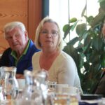 Trije udeleženci – dva moška in ena ženska – poslušajo pogovor. Ženska v sredini govori, moška ob njej zbrano poslušata. V ozadju velika rastlina in svetla steklena stena. Na mizi pred njimi so vrči z vodo in skodelice.
