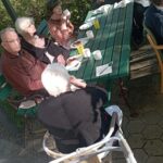 Več starejših stanovalcev sedi okoli zelenega lesenega vrta mize na prostem. V rokah držijo skodelice in kruhke, na mizi so krožniki z namazi in prtički. Jutranje sonce osvetljuje prizor v zelenem vrtu.