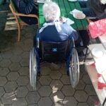 Pogled na zunanjo vrtno mizo, za katero več stanovalcev, tudi v invalidskem vozičku, uživa zajtrk. Na mizi so skodelice, krožniki s kruhom in namazi ter topli napitki.