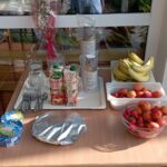 Postrežna miza na prostem z zajtrkom, na kateri so sadje, sokovi, jogurti, voda, plastični kozarci in pribor.