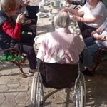 Skupina stanovalcev doma, ki ob jutranjem soncu sedi za zunanjo mizo in zajtrkuje; v ospredju oseba v invalidskem vozičku.