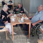 Stanovalci doma za starejše ob pogrnjeni zunanji mizi z zajtrkom; nekateri sedijo v invalidskih vozičkih.