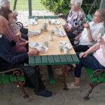 Stanovalci doma med zajtrkom na prostem, obkroženi z zelenjem in prijetnim vzdušjem pod pergolo.