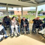 Skupina sedmih starejših ljudi, večinoma v invalidskih vozičkih, sedi pod belo pergolo. Osebe so nasmejane in sproščene, eden od moških salutira. V ozadju je moderno dvorišče z zelenico in parkiriščem.