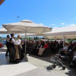 Skupina ljudi v tradicionalnih slovenskih nošah pleše folklorni ples na prostem pred domom za starejše občane. Občinstvo, večinoma starejši v invalidskih vozičkih ali na stolih, opazuje predstavo pod velikimi senčniki in šotorom.