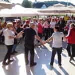 Skupina ljudi, vključno s stanovalci doma starejših, prostovoljci in zaposlenimi, pleše v krogu na ploščadi pred domom. Občinstvo jih spremlja izza senčnikov, ozadje razkriva podeželsko okolje s cerkvico.