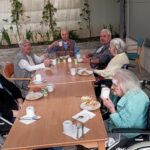 Skupina stanovalcev doma za starejše sedi za mizo zunaj, pijejo kavo in jedo zajtrk v prijetnem dopoldanskem soncu.
