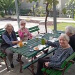 Štiri stanovalke doma za starejše sedijo za zeleno leseno mizo na prostem in uživajo zajtrk.