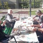 Stanovalci sedijo ob zunanji mizi, pogovarjajo se in uživajo zajtrk v sončnem vremenu.