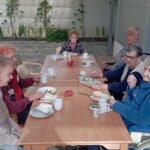 Več stanovalcev, med njimi tudi osebe na invalidskih vozičkih, sedi ob dolgi mizi in zajtrkuje.