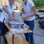 Dve nasmejani zaposleni v belem strežeta sladoled zunaj doma za starejše občane, z ozadjem stavb in zelenega hriba.