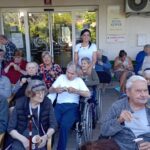 Stanovalci doma za starejše jedo sladoled pred vhodom doma, ob njih stoji zaposlena v belem.