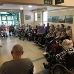 Starejša ženska stoji in govori pred skupino stanovalcev doma starejših, ki sedijo v polkrogu v skupnem prostoru. Nekateri so na invalidskih vozičkih, drugi uporabljajo hodulje.
