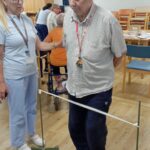 Starejši moški hodi ob pomoči terapevtke, ki ga spremlja, med vadbo hoje čez oviro v obliki napete vrvice.
